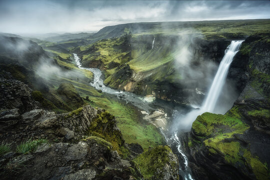 View of Haifoss waterfall cascading into a deep canyon with a winding river and misty green hills, Skeida- og Gnupverjah, Iceland.