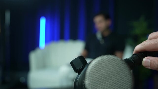 Close-up of professional microphone in a podcast studio with host adjusting the stand. Blurred guest sitting on sofa in the background, media broadcasting and recording concept