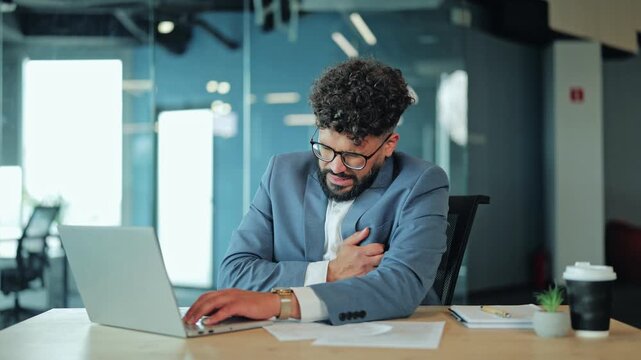 Overworked, pained Middle Eastern executive sits at office desk, clutching chest. Tired professional experiencing severe heart discomfort, physical stress, and sudden health crisis during work hours.