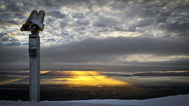 View of Col de la Faucille with a yellow binocular telescope overlooking golden sunbeams breaking through clouds over the valley in Gex, Auvergne-Rhone-Alpes, France.