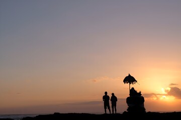 Fototapeta premium Minimalist silhouette of two people and traditional Balinese shrine with ceremonial umbrella against clean golden orange dusk sky over Bali ocean.