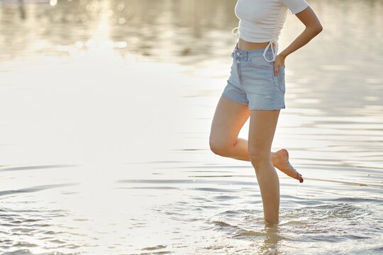 Unrecognizable woman steps carefully in the clear water, enjoying the warmth of the sun on her skin. Wearing a light top and denim shorts, she experiences the refreshing sensation of the water