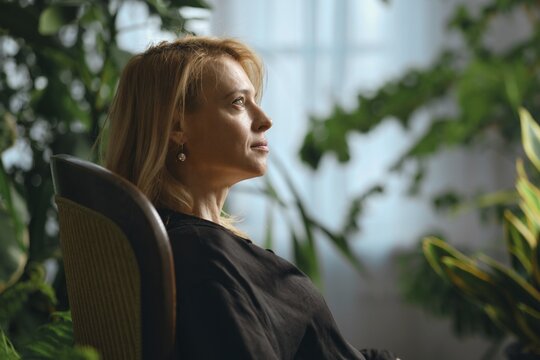 A woman with long blond hair sits thoughtfully in a wooden chair, surrounded by vibrant green plants in a well-lit room. Sunlight illuminates her face as she gazes into the distance
