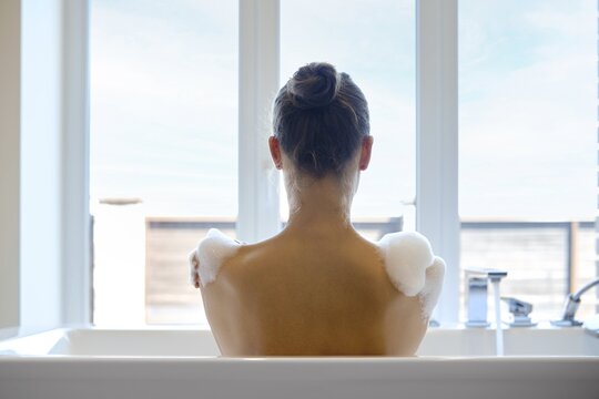 Unrecognizable woman enjoys a calming bath in a modern tub. A soap foam is gently resting on her shoulders. Bright natural light filters through large windows, creating a tranquil atmosphere for relaxation