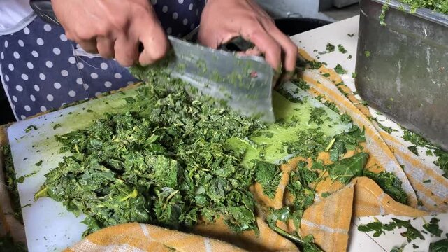 A person is chopping boiled cassava leaves with a large knife on a wooden cutting board. commonly served with traditional cuisine from East Nusa Tenggara (NTT), Medan, Sulawesi, and Papua.