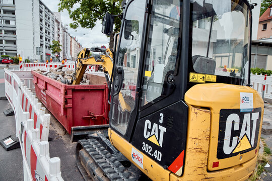 A yellow Caterpillar 302.4D mini excavator working at an urban road construction site next to a large red waste skip container.