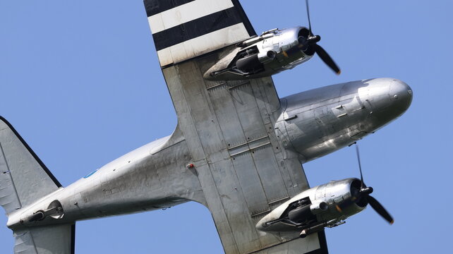 View of the underbelly of a vintage silver propeller aircraft with invasion stripes flying against a clear blue sky in La Ferte-Alais, Ile-de-France, France.