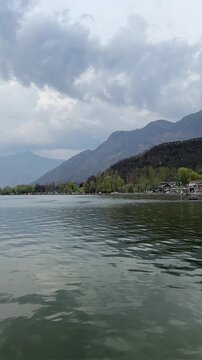 A scenic boat-level view of Dal Lake, Srinagar, Kashmir, with rippling green waters, lush tree-lined shores, traditional houseboats, and the majestic Zabarwan mountains beneath a dramatic cloudy sky.