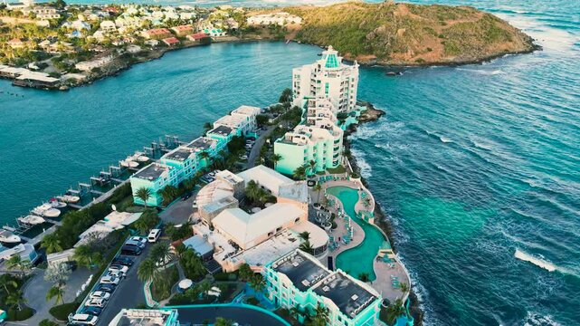 Drone view of the luxury beach resort situated on a peninsula. The modern hotel buildings stand on the coastline facing the Caribbean Sea. This location featuring a large curved swimming pool.
