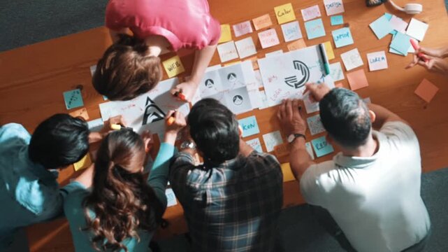 Top down aerial view of skilled designer hold logo while discuss with team. Aerial business group working together sharing idea about logo graphic design by using sticky notes and tablet. Symposium.