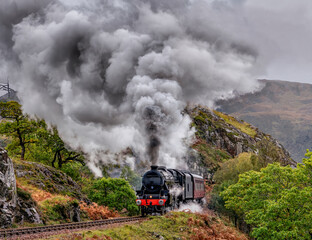 Naklejka premium Steam train emerging from a tunnel in the Scottish Highlands, Scotland. Dramatic clouds of steam and smoke create a classic railway scene.