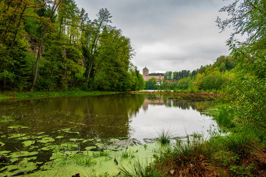 Kost Castle rises above a pond in the Bohemian Paradise region of the Czech Republic. Forests surround the landscape under cloudy skies, creating a scenic historic view.