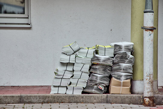 A large stack of bundled newspapers and advertising flyers delivered and piled up on a sidewalk next to a building wall and a pipe.