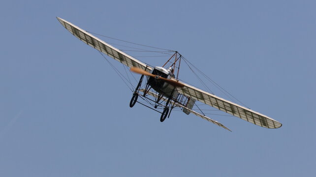 View of a vintage monoplane with a wooden propeller and wire-braced wings flying through a clear blue sky during an airshow in La Ferte-Alais, Ile-de-France, France.