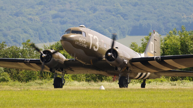 View of a vintage Douglas C-47 Skytrain military transport aircraft taxiing on a grassy airfield with lush green hills in the background La Ferte-Alais, Ile-de-France, France.