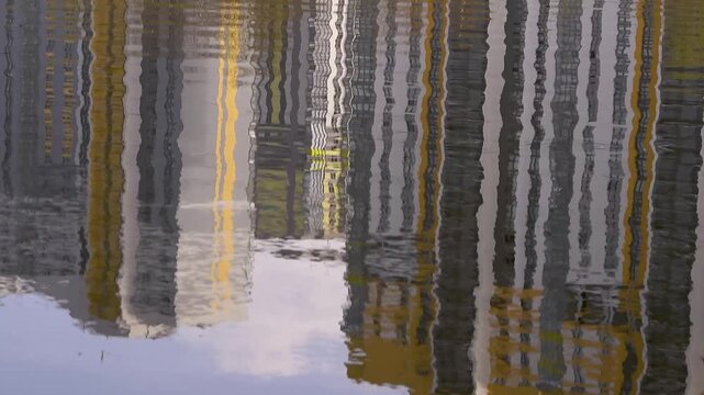 Vertical reflections of tall city buildings ripple across a calm water surface, creating abstract dark and light bands with soft sky tones near the edge