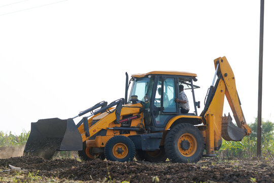 A yellow backhoe loader is digging and leveling soil in a rural agricultural field
