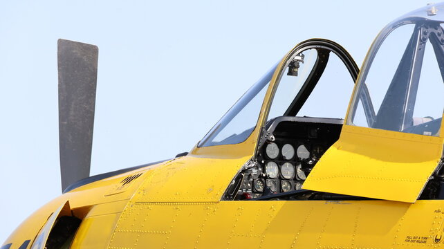 View of a yellow vintage aircraft cockpit with an open canopy, detailed instrument panel, and propeller blade under a clear sky in La Ferte-Alais, Ile-de-France, France.