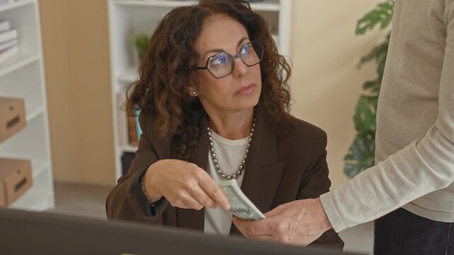 Middle age woman takes cash from a man's hand while seated at a desk in a building; bribery tension.