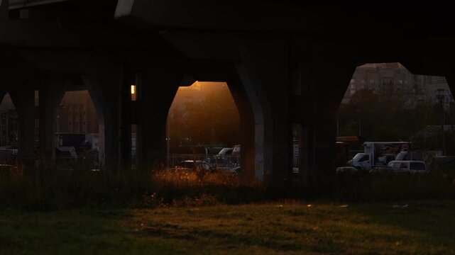 Dim urban underpass with large concrete arches, a warm orange glow in the center opening, grass in the foreground, and distant buildings and parked vehicles