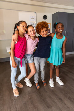 Four children standing with arms around, posing in classroom near whiteboard clock grey cabinet