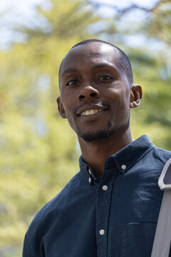 African adult man smiling and looking at camera in park, wearing navy shirt, shoulder bag strap