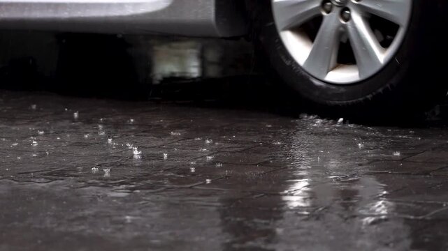 Close-up low-angle view of a moving car wheel crossing a wet roadway in rainy low light, with water splashing around the tire and reflections shimmering on