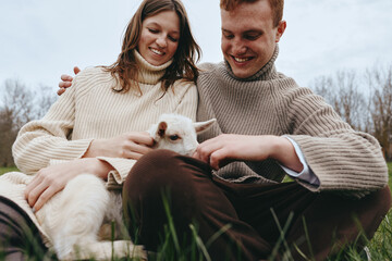 Naklejka premium Cheerful couple outdoors, gently patting a curious goat kid, knit sweaters, sunlit grass, calm atmosphere, natural colors, candid moment, joyful connection, rural setting
