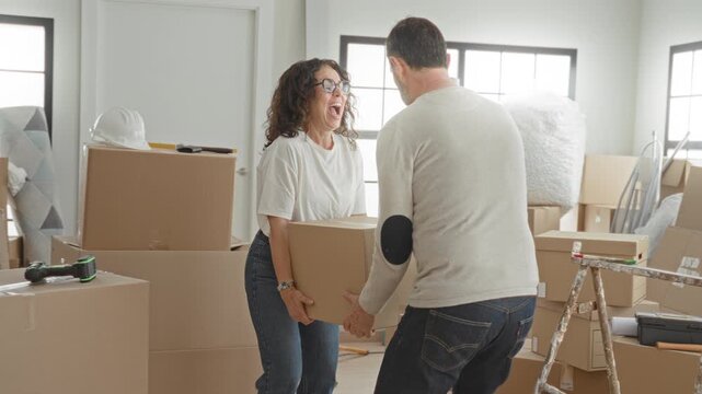 Man and woman hold cardboard box with hands while smiling amid packed moving boxes, ladder and toolbox in sunlit building; teamwork joy.