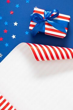 Vertical layout of Fourth of July gift box with blue ribbon bow on blue background with star confetti and unrolled striped wrapping paper. 4th of July Independence Day patriotic backdrop with copy spa