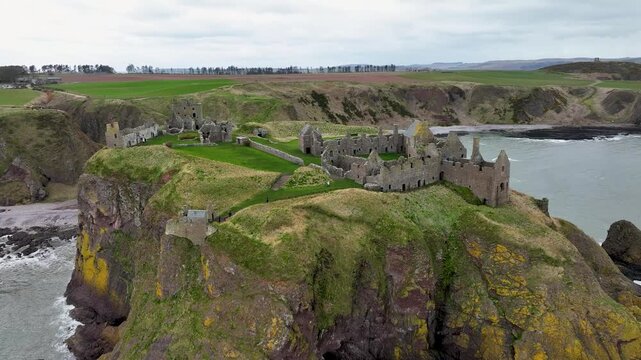 Aerial view of Dunnottar Castle ruins perched dramatically on a cliffside with crashing waves below and green fields behind, Stonehaven, Scotland.