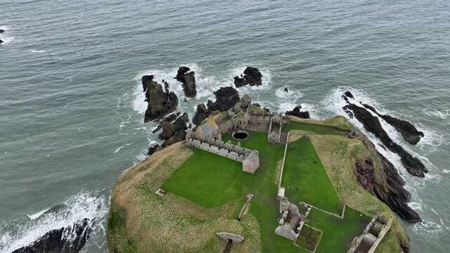 Aerial view of Dunnottar Castle ruins perched on a dramatic cliff edge, where the green grass meets the rugged coastline and turbulent sea, Stonehaven, Scotland.