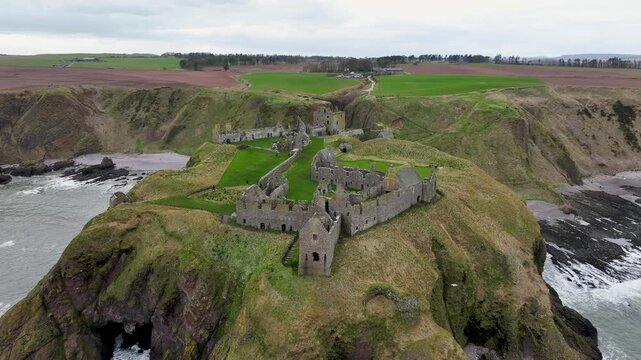 Aerial view of the historic Dunnottar Castle ruins perched on a rugged cliff, contrasting with the green grass and crashing waves, Stonehaven, Scotland.