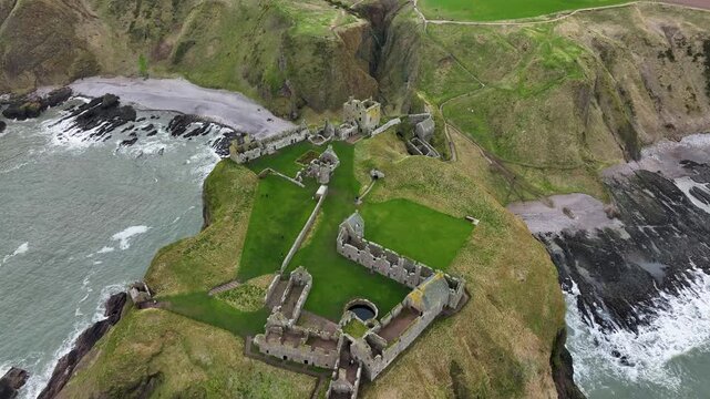 Aerial view of Dunnottar Castle ruins, perched atop verdant cliffs meeting the turbulent sea, a stark contrast of green grass and grey stone, Stonehaven, Scotland.