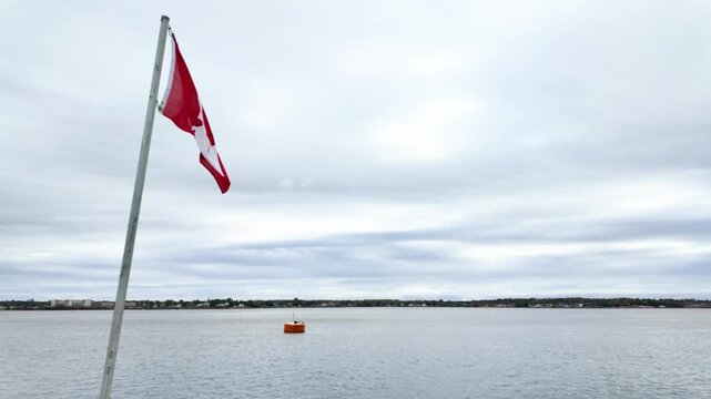 Canadian flag flying above a calm harbour in Nova Scotia Canada with coastal shoreline distant waterfront buildings and overcast sky creating a peaceful maritime scene beside sheltered waters