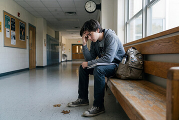 Fototapeta premium A young student with dark hair sits on a bench in a school hallway. He covers his face with his hand, looking down. His bag rests on the floor, and leaves are scattered around