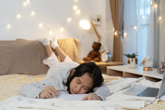 Young girl studying on bed in bedroom warm lighting with notebooks and a laptop, drifting to sleep during study session