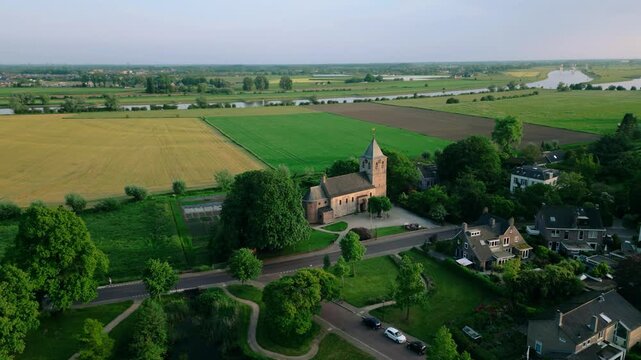 Aerial view of the historic Oude Kerk in Oosterbeek, Netherlands, showcasing its ancient architecture amidst lush green fields and trees under a serene blue sky. Captures the tranquil village setti...