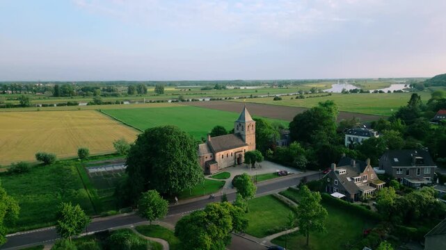 Aerial view of the historic Oude Kerk in Oosterbeek, Netherlands, showcasing its ancient architecture amidst lush green fields and trees under a serene blue sky. Captures the tranquil village setti...