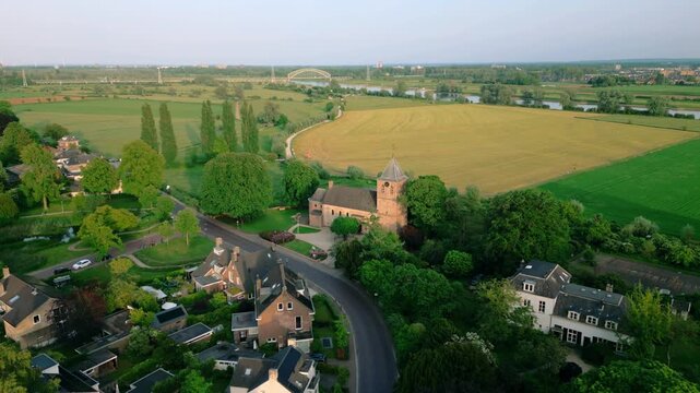 Aerial view of the historic Oude Kerk in Oosterbeek, Netherlands, showcasing its ancient architecture amidst lush green fields and trees under a serene blue sky. Captures the tranquil village setti...