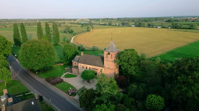 Aerial view of the historic Oude Kerk in Oosterbeek, Netherlands, showcasing its ancient architecture amidst lush green fields and trees under a serene blue sky. Captures the tranquil village setti...