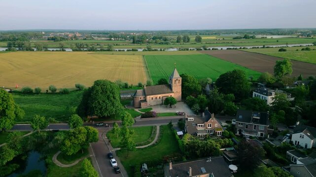 Aerial view of the historic Oude Kerk in Oosterbeek, Netherlands, showcasing its ancient architecture amidst lush green fields and trees under a serene blue sky. Captures the tranquil village setti...