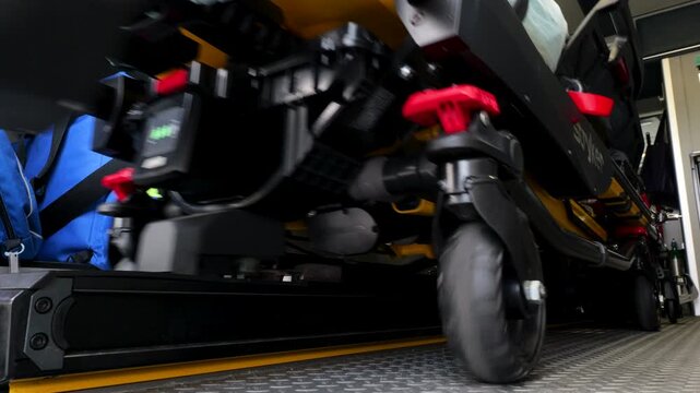 Motorized ambulance stretcher with powered wheel system being unloaded from emergency vehicle platform, showing medical transport equipment and paramedic handling procedures, static low angle closeup