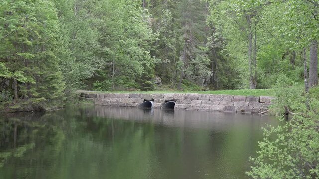 Low stone dam wall spanning across calm water with two black semi-circular culvert pipes. Dense mixed forest rises behind the structure.