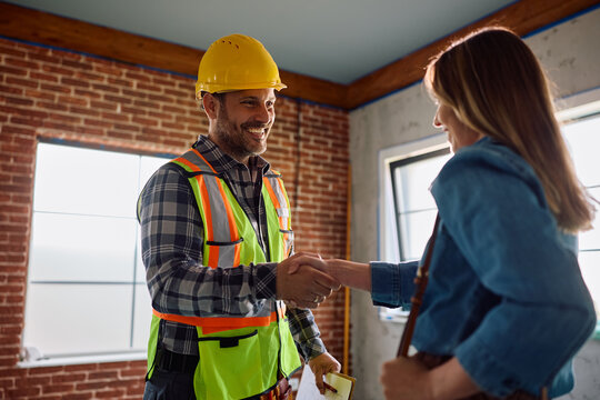 Happy construction worker and female homeowner shaking hands on site.