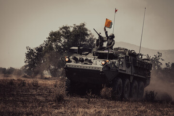 Naklejka premium Dramatic shot of an eight-wheeled green military armored vehicle (APC) advancing through a dense dust and haze cloud on a dry dirt field under heavy golden sunset light.