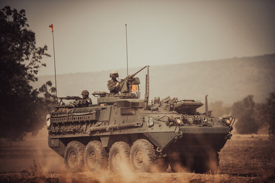 Dramatic shot of an eight-wheeled green military armored vehicle (APC) advancing through a dense dust and haze cloud on a dry dirt field under heavy golden sunset light.