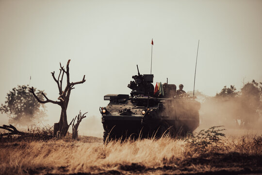 Dramatic shot of an eight-wheeled green military armored vehicle (APC) advancing through a dense dust and haze cloud on a dry dirt field under heavy golden sunset light.