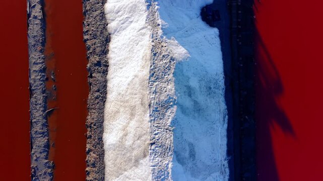 Overhead close-up of salt mound between crimson brine pools, Bulgaria. Aerial view of white salt ridge flanked by deep red mineral water and dark levee, tire tracks visible.