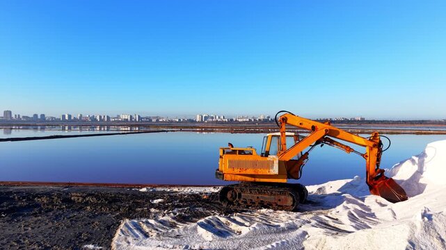 Yellow excavator harvesting salt beside evaporation ponds, Bulgaria. Aerial side view of orange tracked machine next to white salt pile, reflective blue pools and city skyline in background.